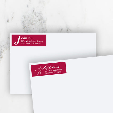 Christmas red address stickers on 2 white envelopes. One with classic serif font and one with modern script font on marble table.