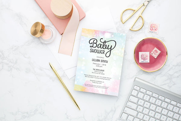 Wide view of rainbow watercolor textured baby shower invitation with gold pen and pink desk accessories.