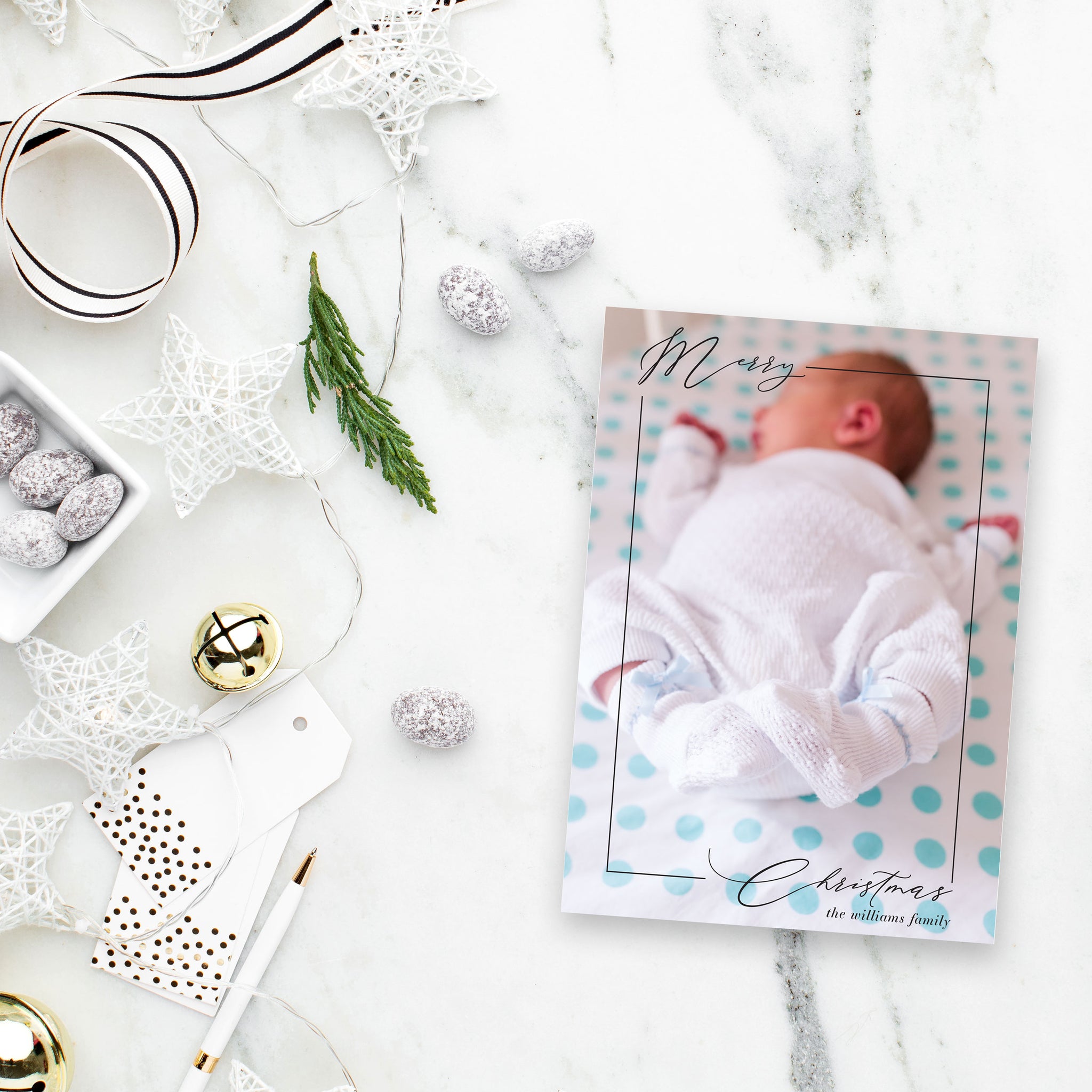 Close up of baby feet with baby sleeping on holiday photo card with thin script "Merry Christmas". White and gold holiday decor surrounds.