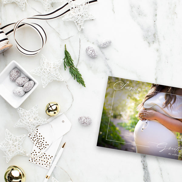 Woman holding pregnant belly on holiday photo card with thin script "Merry Christmas". White and gold holiday decor surrounds.