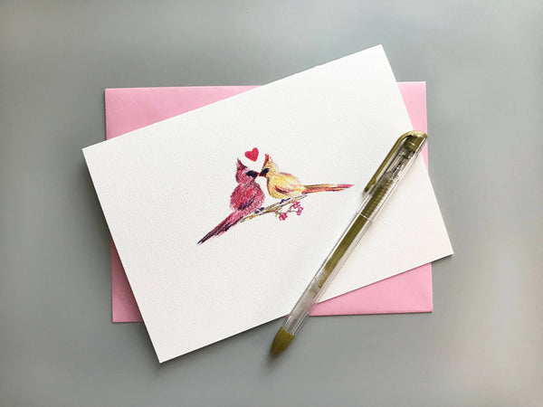 Colored pencil drawing of cardinals kissing with a heart on greeting card with pink envelope and gold ink pen.