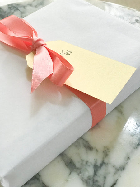 Perspective image of wrapped package with white paper, coral satin ribbon in a bow, and a gold name tag with script "to" and "from". Marble background.