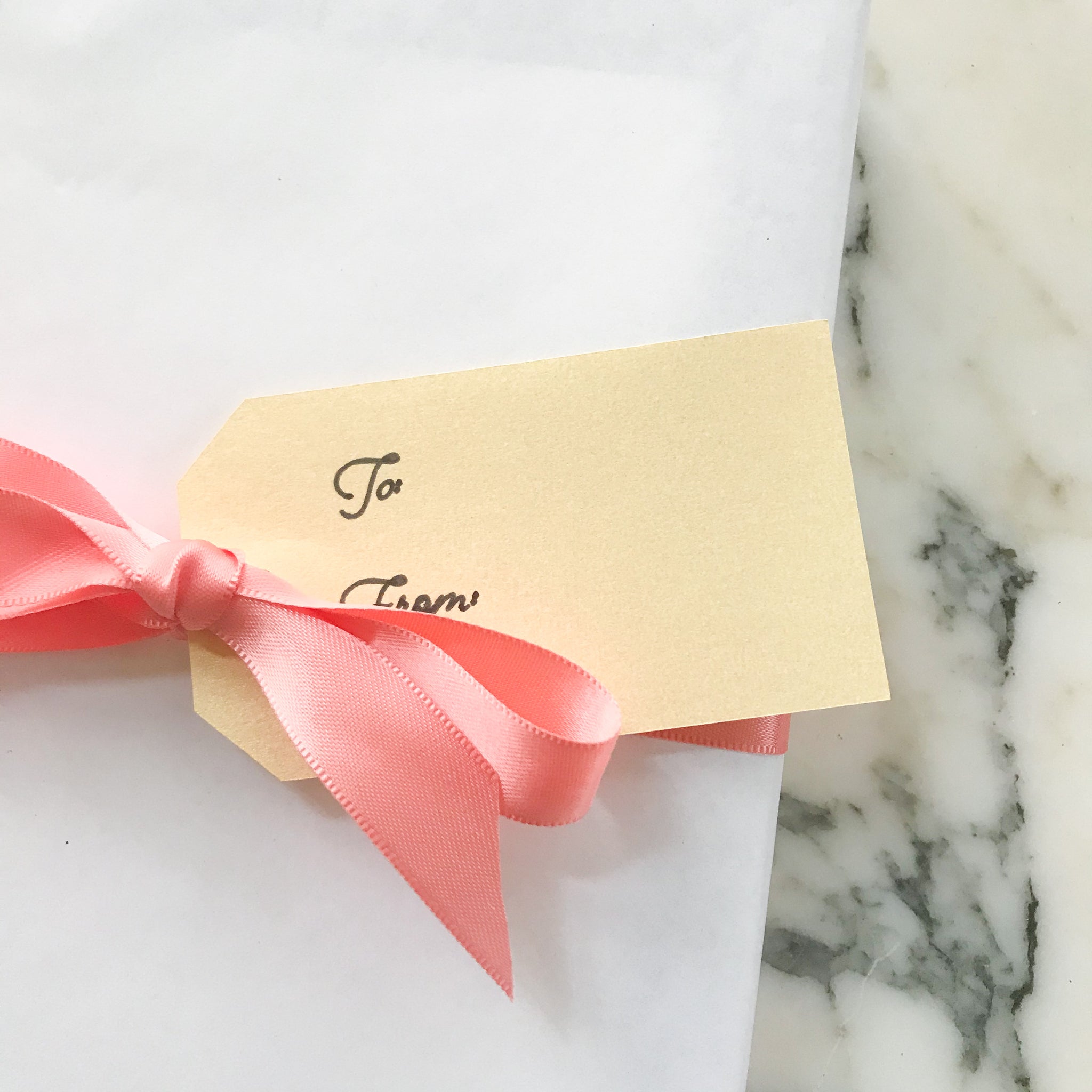 Wrapped package with white paper, coral satin ribbon in a bow, and a gold name tag with script "to" and "from". Marble background.