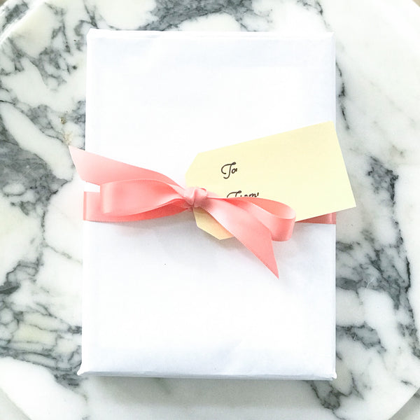 Entire image of wrapped package with white paper, coral satin ribbon in a bow, and a gold name tag with script "to" and "from". Marble background.