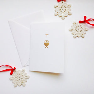 Gold foil card design with baby in manger and star. White ceramic snowflake ornaments with red ribbon surround.