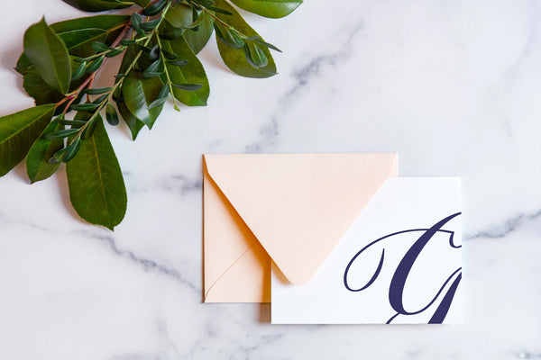 Monogram stationery with single script "G" bleeding off the card with a blush envelope on a marble table top.