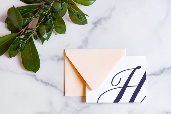 Monogram stationery with single script "H" bleeding off the card with a blush envelope on a marble table top.