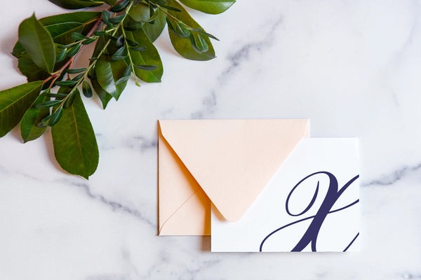 Monogram stationery with single script "X" bleeding off the card with a blush envelope on a marble table top.