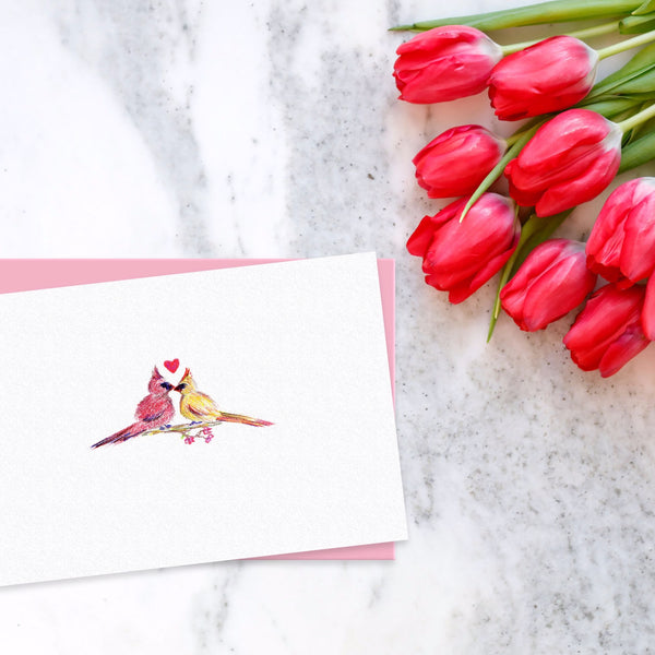 Colored pencil drawing of cardinals kissing with a heart on greeting card surrounded by red tulips on white marble table.