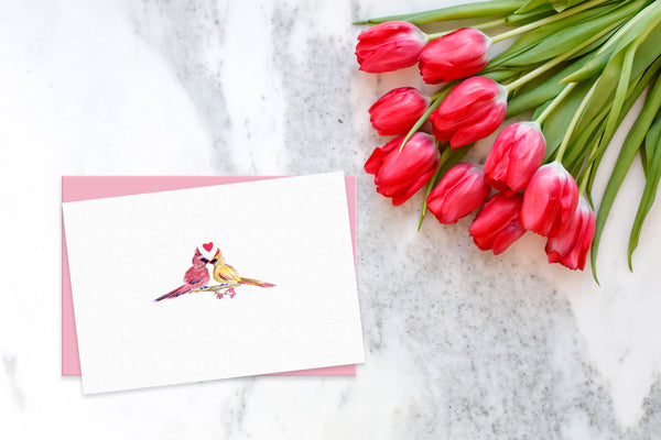 Colored pencil drawing of cardinals kissing with a heart on greeting card surrounded by red tulips on white marble table.