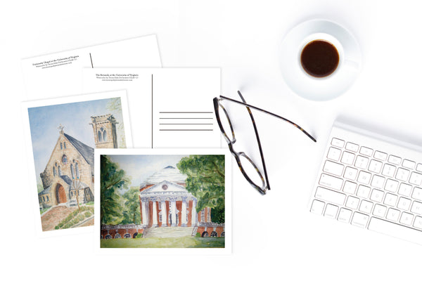 Watercolor postcards of University of Virginia Rotunda and Chapel on white table with keyboard, mug black coffee, and tortoise shell eyeglasses.